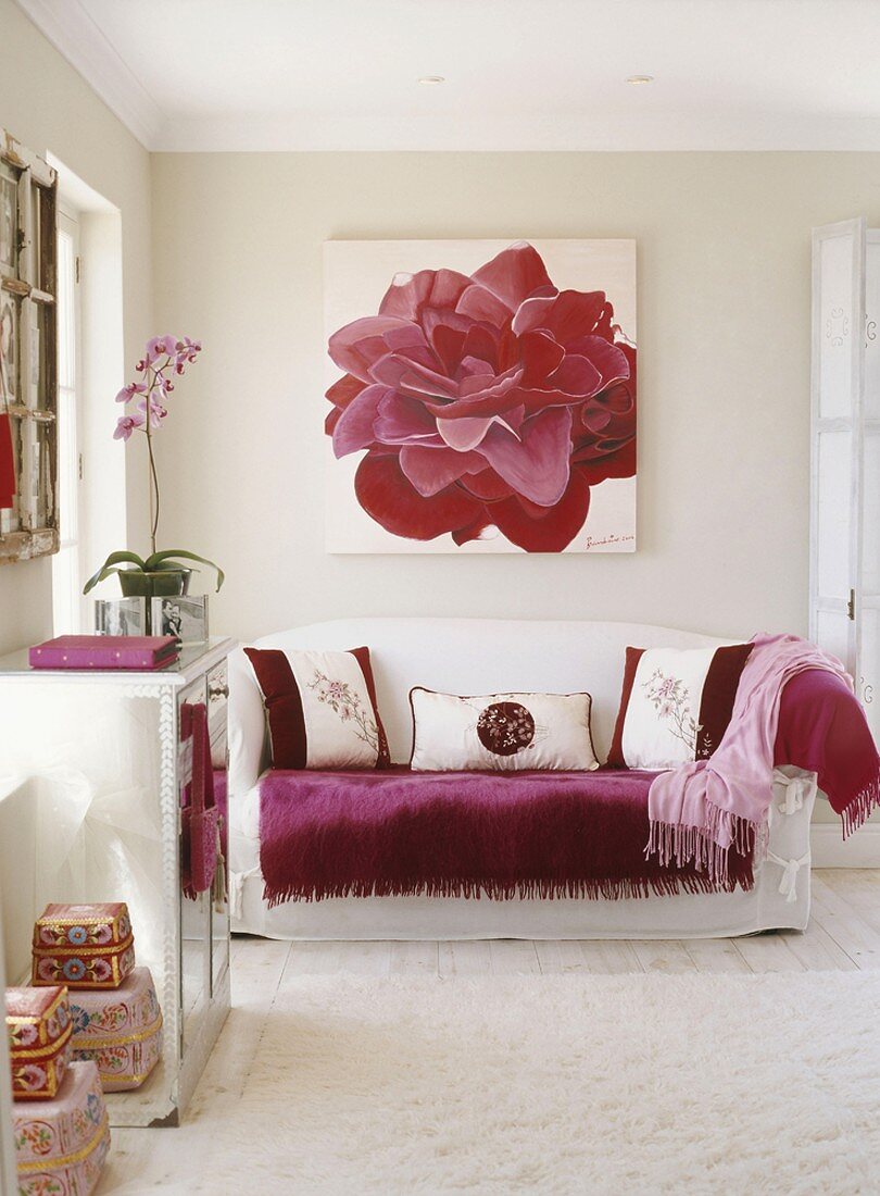 Ein Blumenbild über einer weißen Couch mit bordeaux-roter Decke und rot-weißen Zierkissen im Wohnzimmer