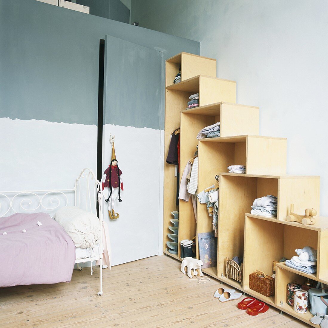 Kinderzimmer mit offenem Kleiderschrank in Stufenoptik, daneben ein weisses Metallbett und eine versteckte Zimmertür