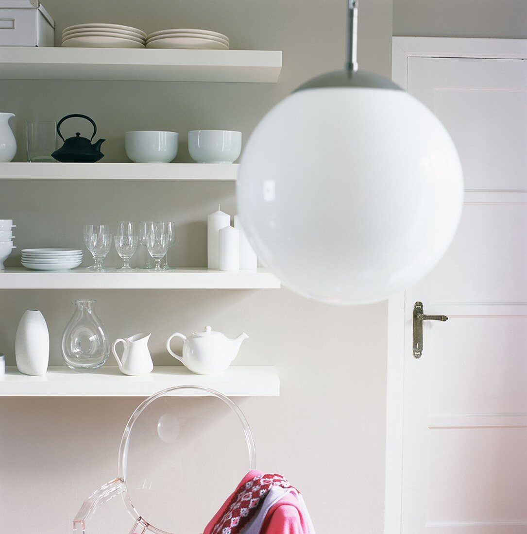 weiße Kugellampe, dahinter ein Wandregal mit Geschirr und ein durchsichtiger Plexiglasstuhl
