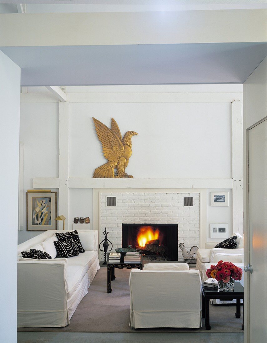 Weisses Wohnzimmer mit weisser Sitzgarnitur, brennendem Kaminfeuer und einer goldenen Adlerfigur auf dem Kaminsims