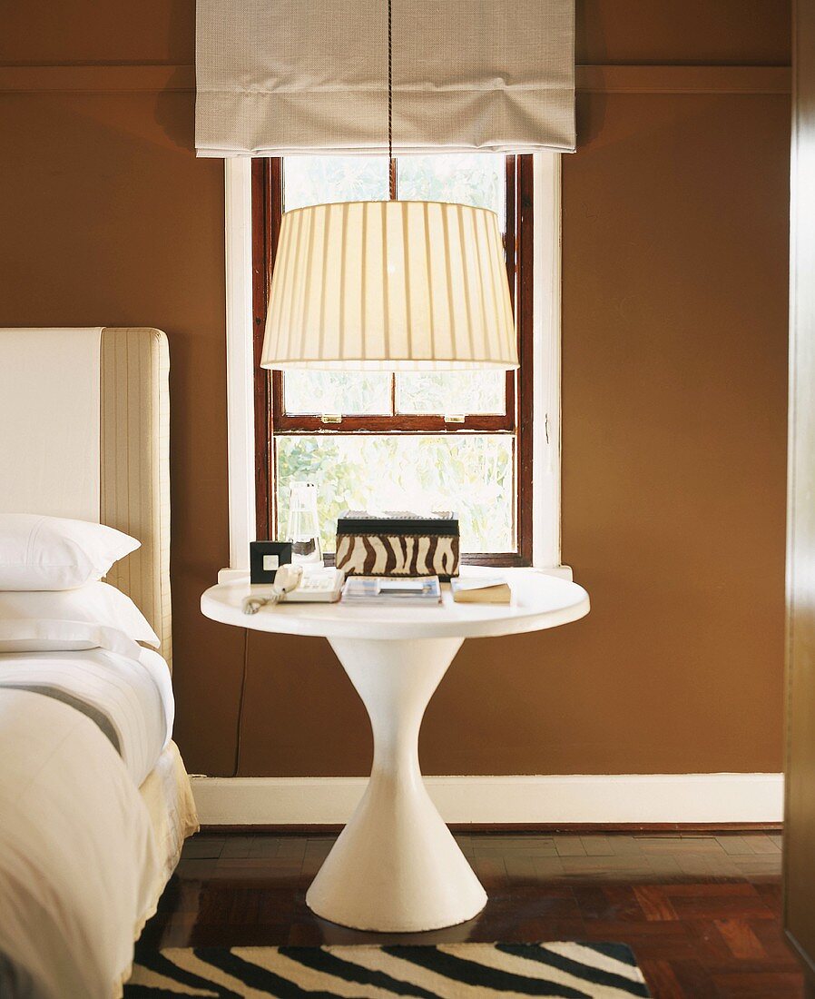 Round, white bedside table below sixties-style lamp next to bed