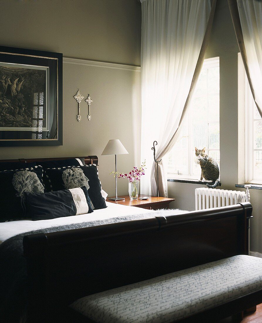 Perlgraues Schlafzimmer mit schwarzem Doppelbett, Kissen mit römischen Kaiserbüsten und mit einer Katze auf der Fensterbank