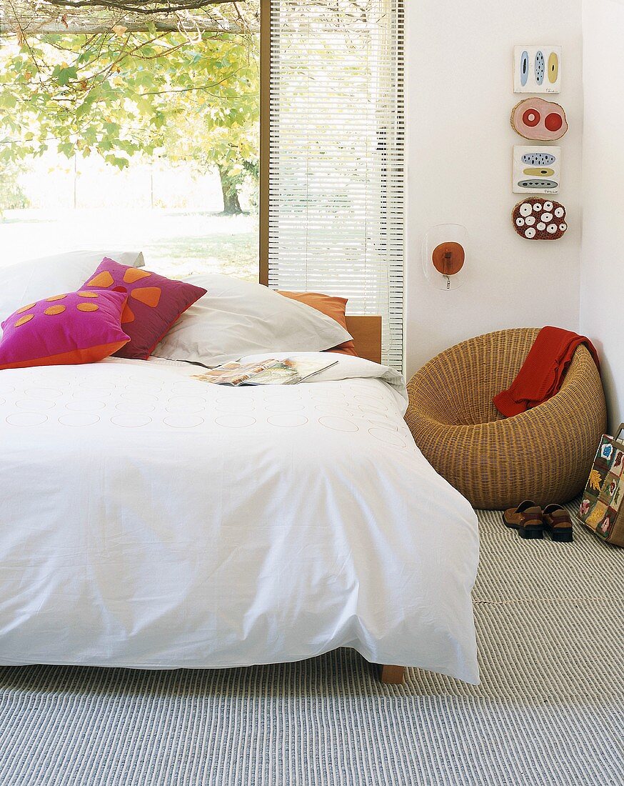 Bett vor offener Terrassentür; in der Ecke ein runder Rattansessel und Kunstobjekte an der Wand