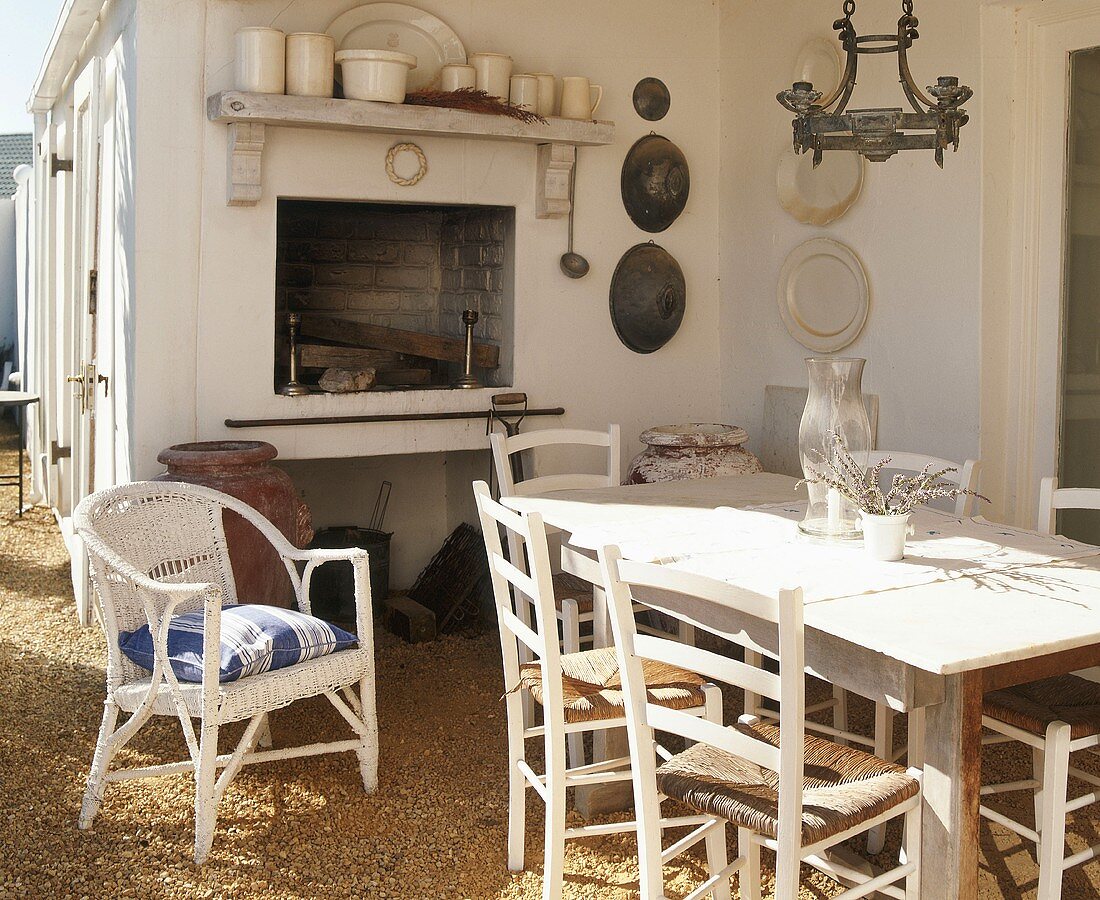 Sunny terrace with open fireplace, rustic dining table, simple rush-seat chairs and one white wicker chair