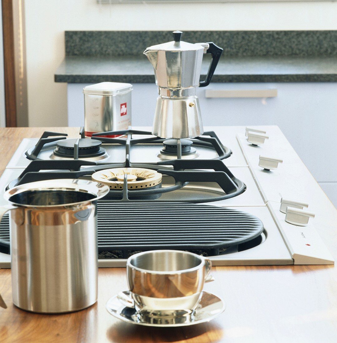 Ein Kaffeekocher auf dem Gasherd, im Vordergrund eine Tasse aus Metall und eine Milchkanne