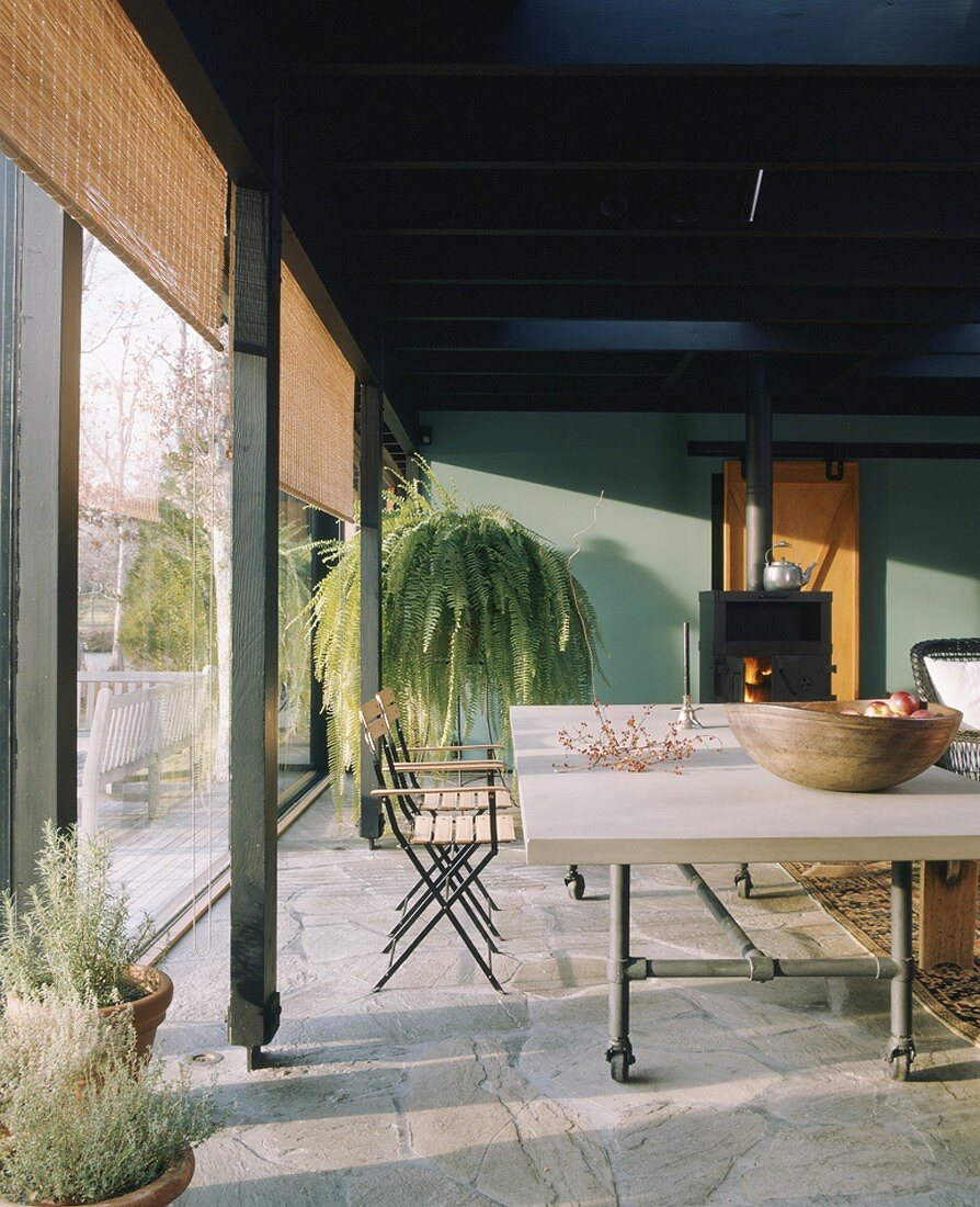 Schwerer Esstisch mit Rollen vor großer Fensterfront mit Bambusrollos; im Hintergrund ein schwarzer Kaminofen und eine üppige Zimmerpflanze