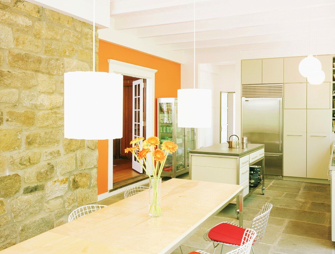 Sonniger Essraum mit Natursteinwand, langer Esstafel mit Metallstühlen, Hängeleuchten und einer zweiten, leuchtendorangen Wand