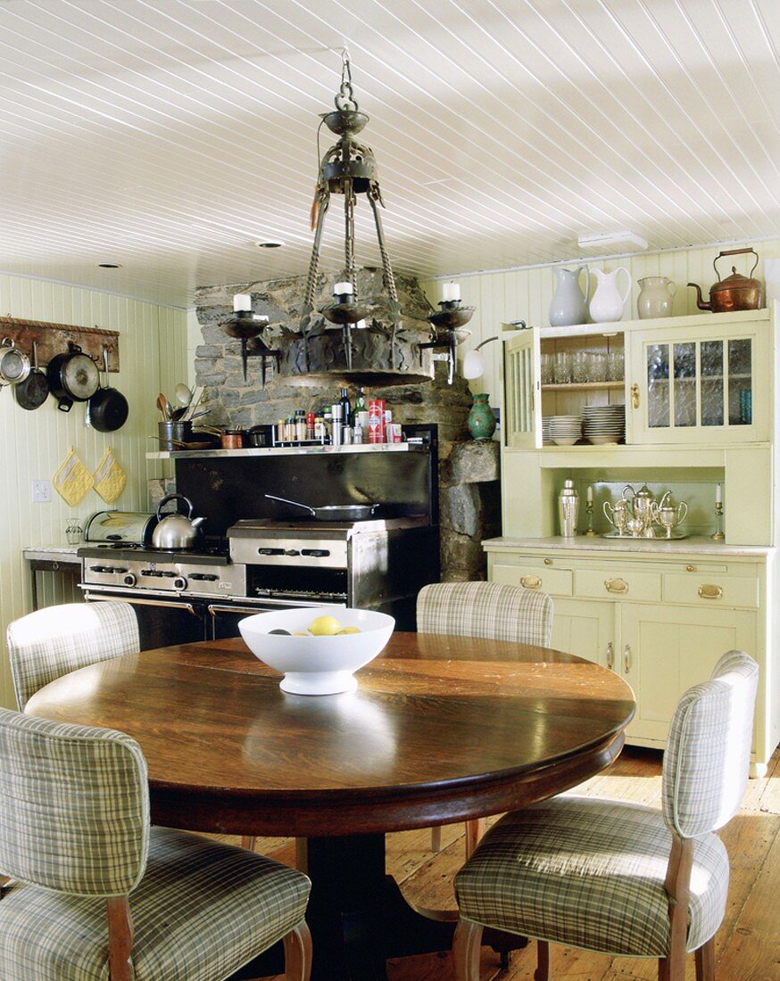 Küche mit Natursteinwand und modernen Küchengeräten, einem weiss lackierten, altmodischen Küchenbuffet und einem runden, robusten Esstisch aus dunklem Holz