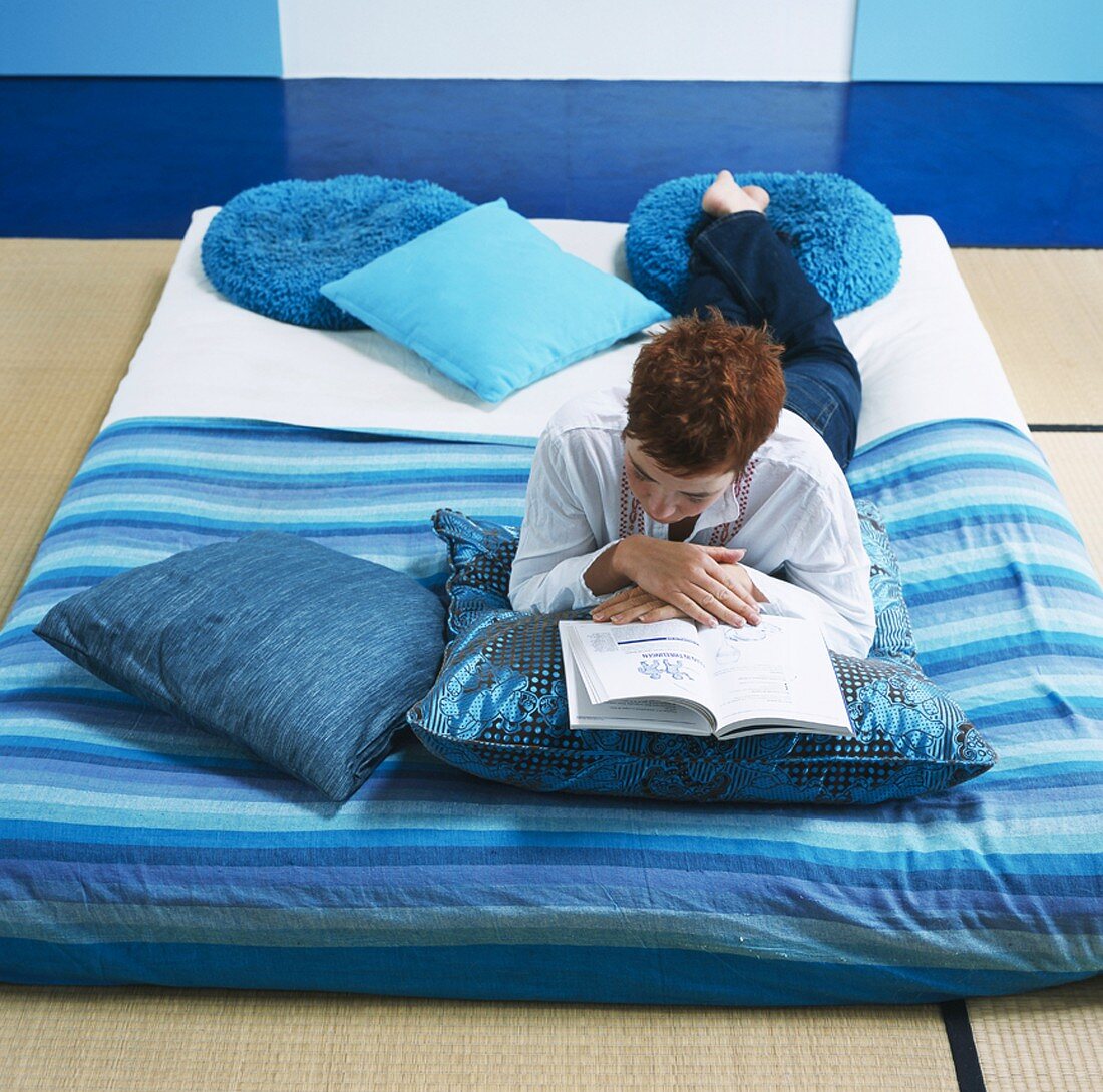 Woman lying reading on futon with blue scatter cushions and blue blanket