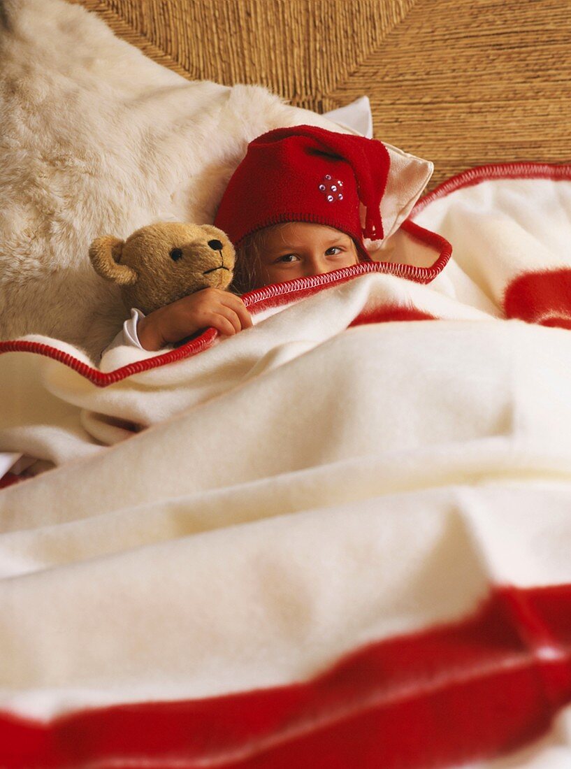 Girl wearing woolly hat and teddy bear under woollen blanket
