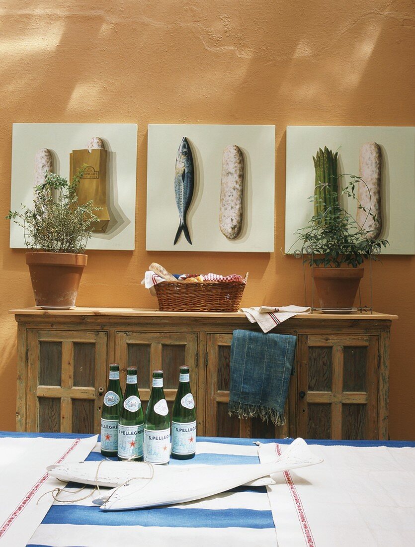 Rustikales Sideboard und Bildserie mit Lebensmitteln, im Vordergrund 4 Wasserflaschen auf einem Tisch