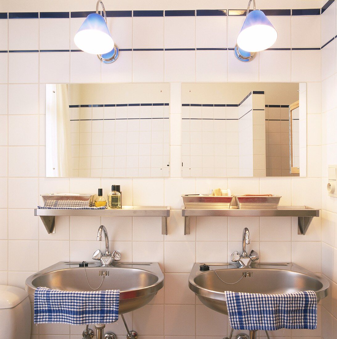 Zwei Edelstahlwaschbecken in einem weiss geflieseten Badezimmer mit blauen Zierleisten
