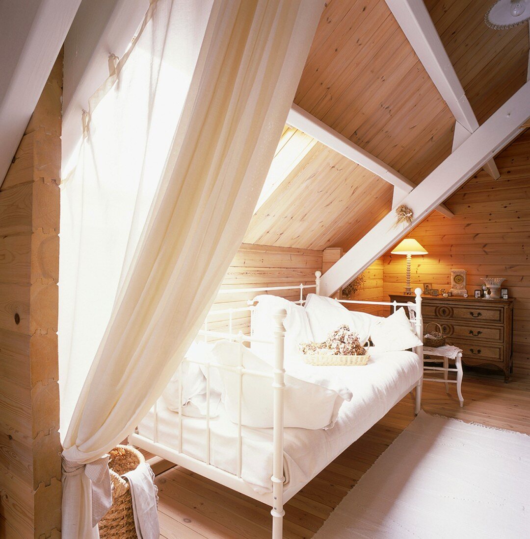 Der holzverkleidete Dachraum eines Wohnhauses dient als Schlafzimmer mit antiker Kommode und einem romantischen Metallbett