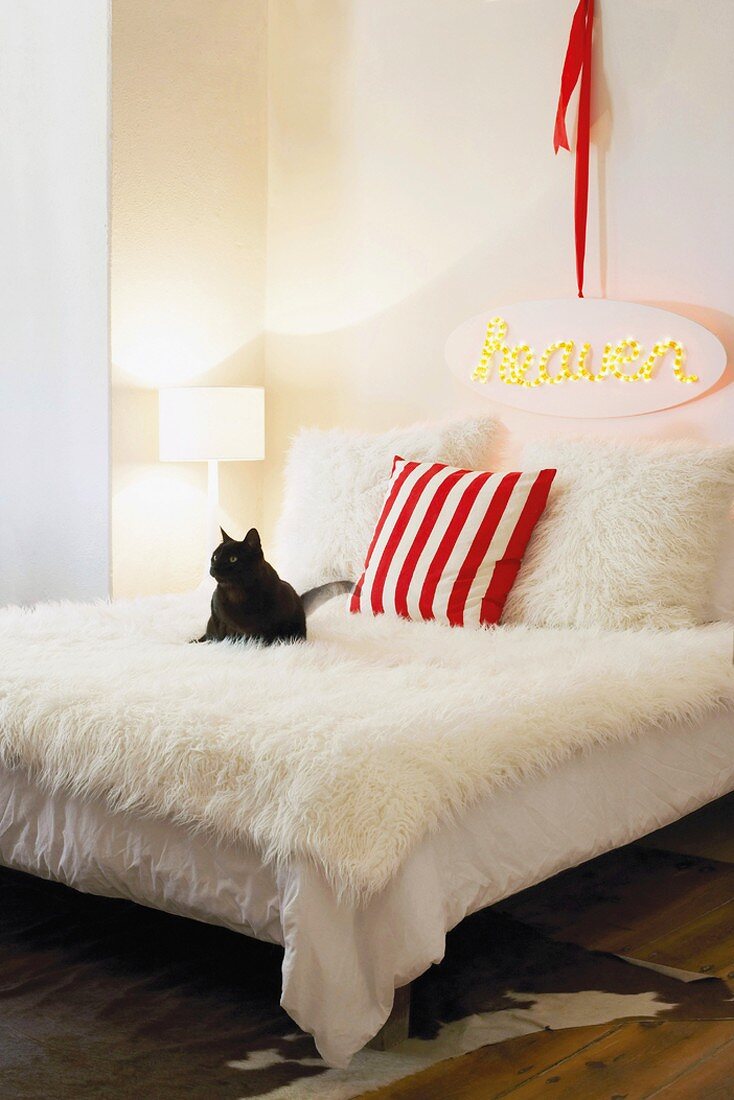 Eine schwarze Katze auf einem poppigen Bett mit weisser Felldecke und -kissen