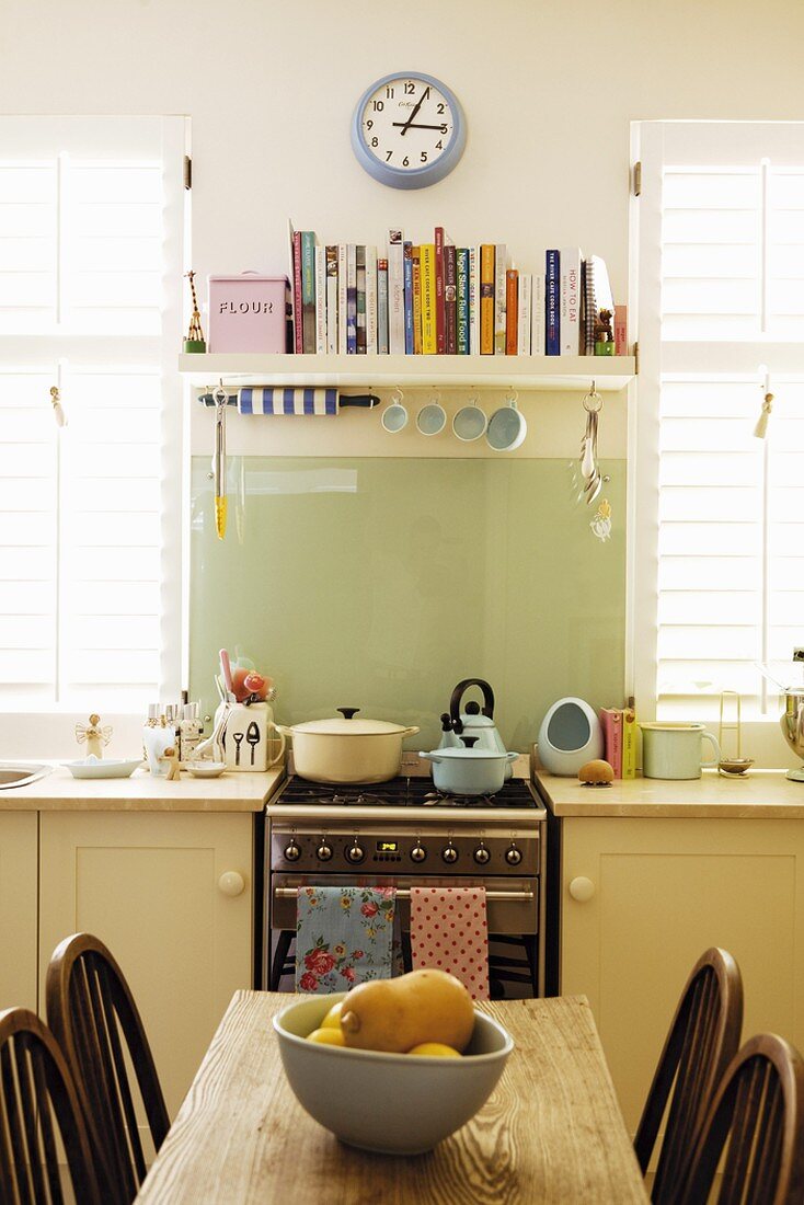Blick über einen rustikalen Holztisch zum Edelstahlgasherd der Küchenzeile und einem einfachen Wandboard mit Kochbüchern