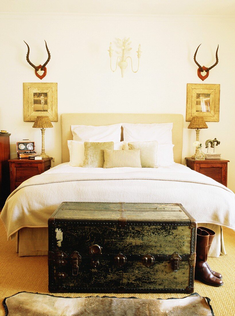 Ein antiker Überseekoffer vor einem gemachten Bett im rustikal dekorierten Schlafzimmer