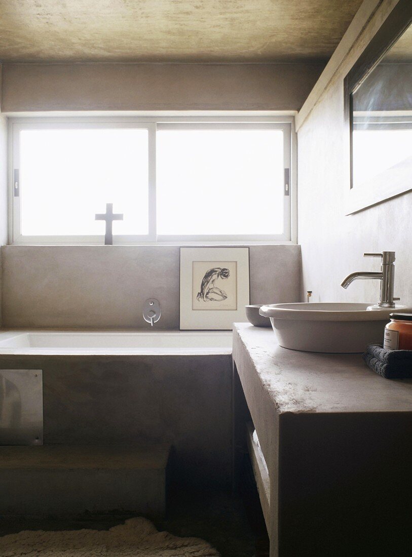 Ein rundum verputztes Badezimmer in dezentem Grau mit einem Langfenster über der Badewanne