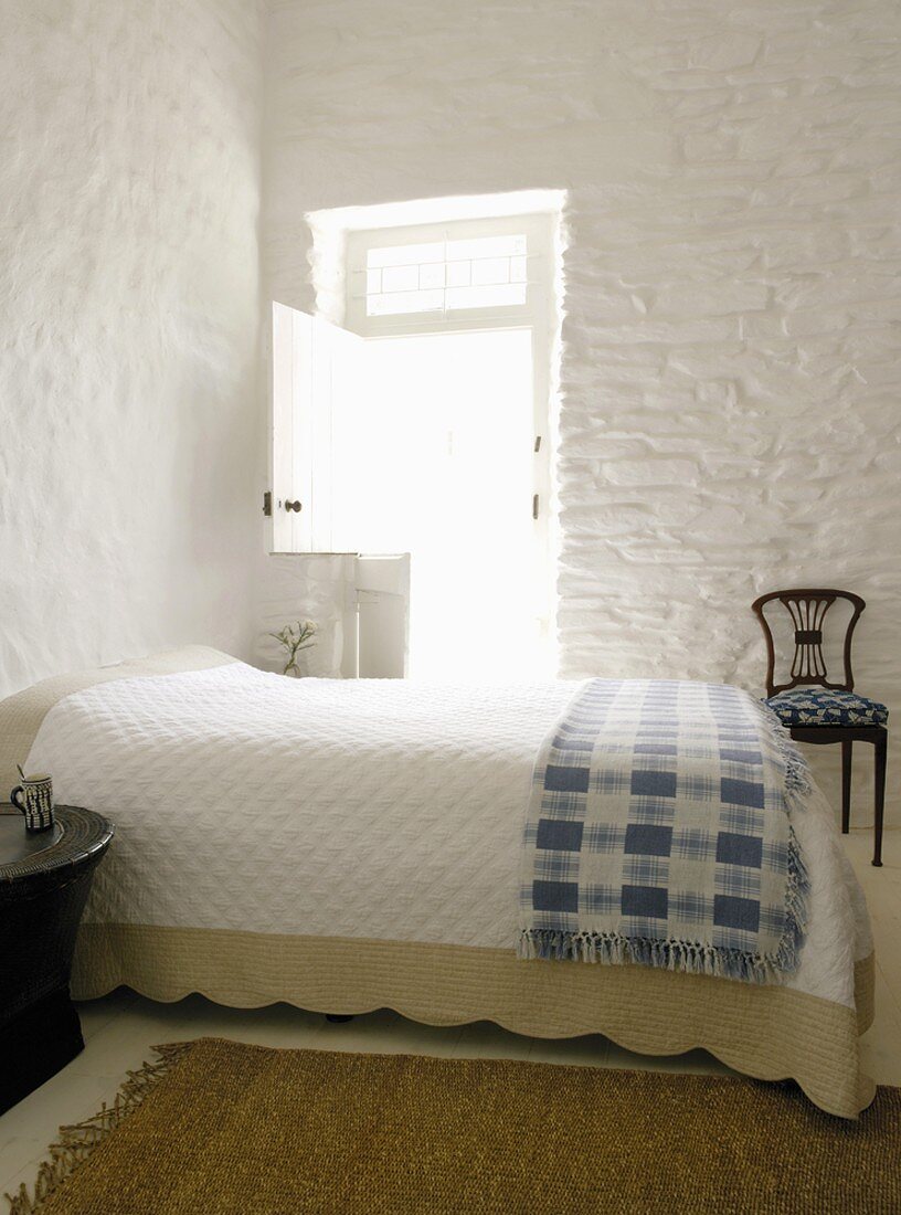 Doppelbett mit Tagesdecke im Schlafzimmer mit weissen Steinwänden und geöffneter Terrassentür