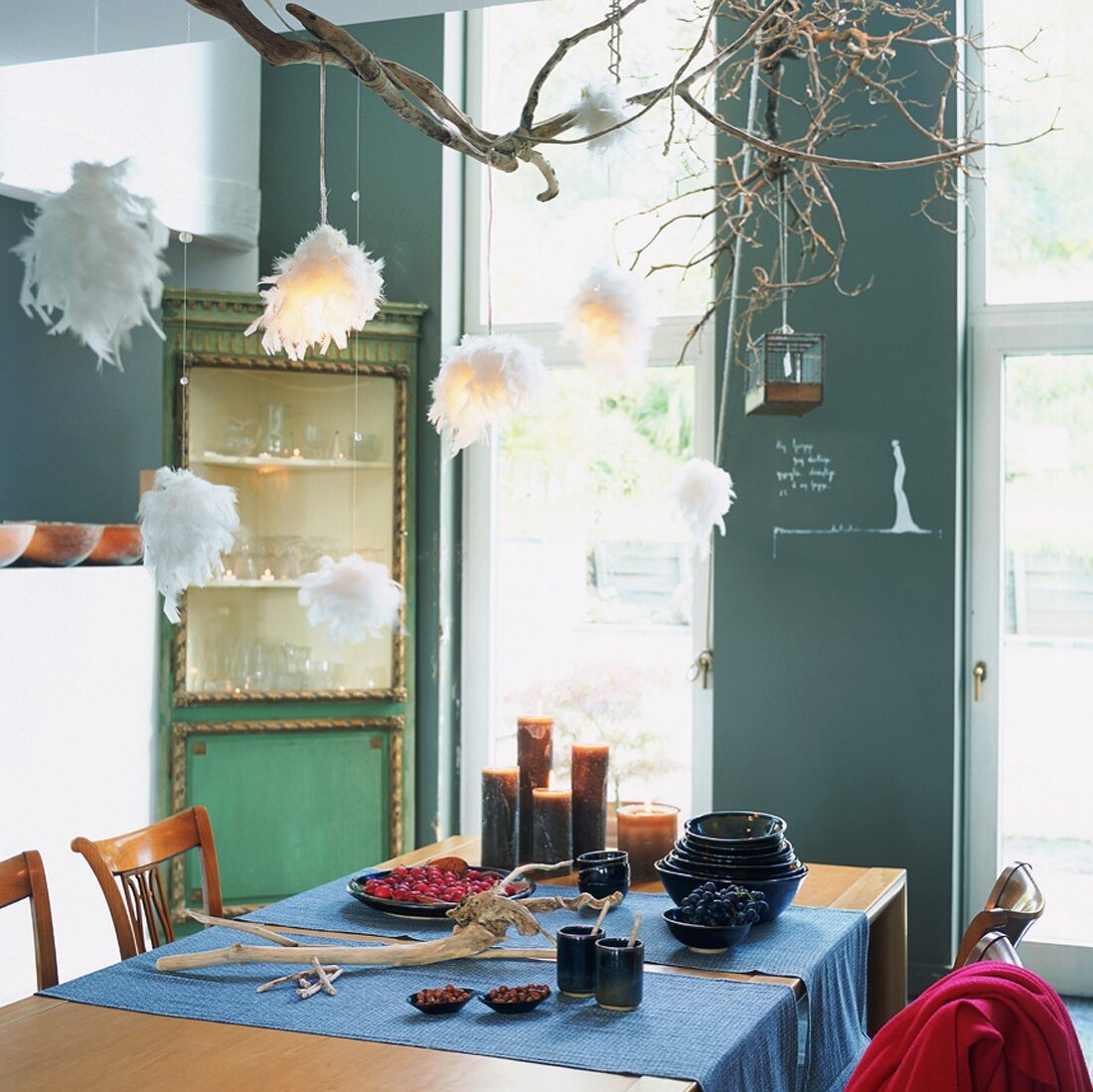 Eine selbstgebaute Hängeleuchte aus Zweigen und Federbällen über einem Esstisch mit Läufern, Kerzen und Tonschalen