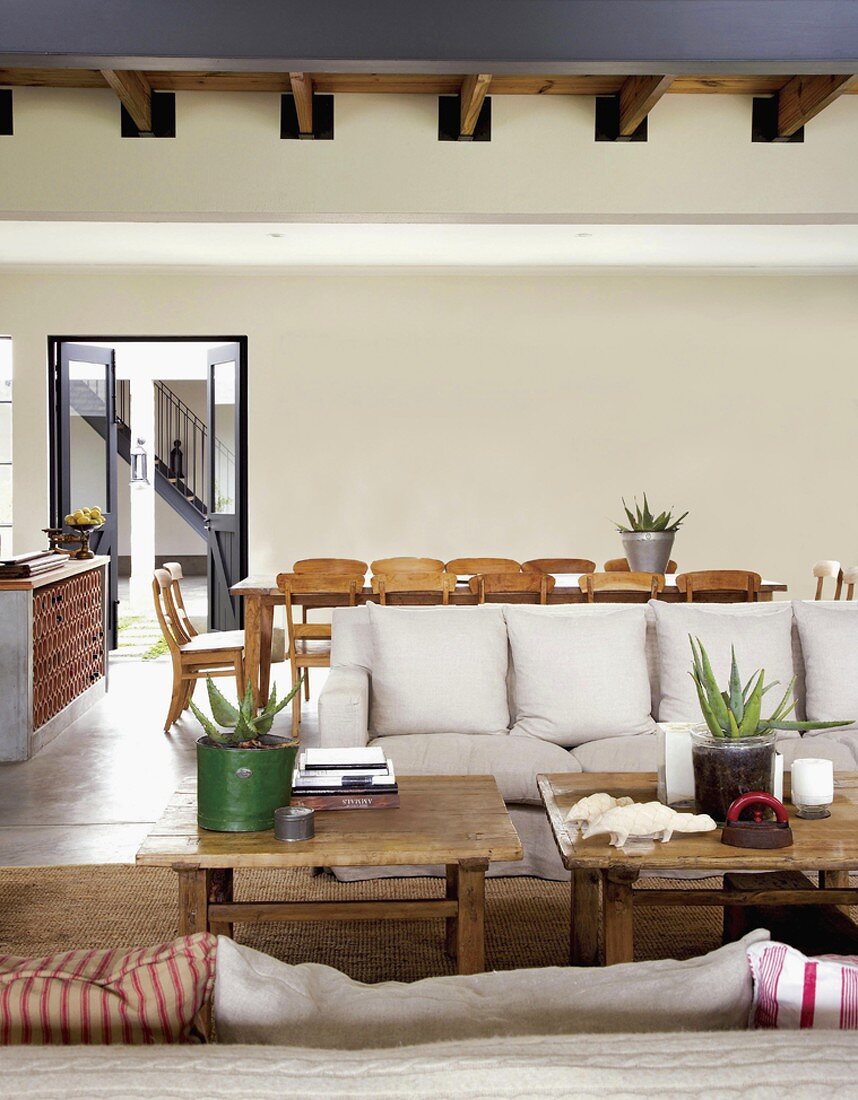Rustikale Couchtische und helle Sofas vor einem großen Esstisch mit Biedermeierstühlen in einem offenen Wohnraum mit Holzbalkendecke