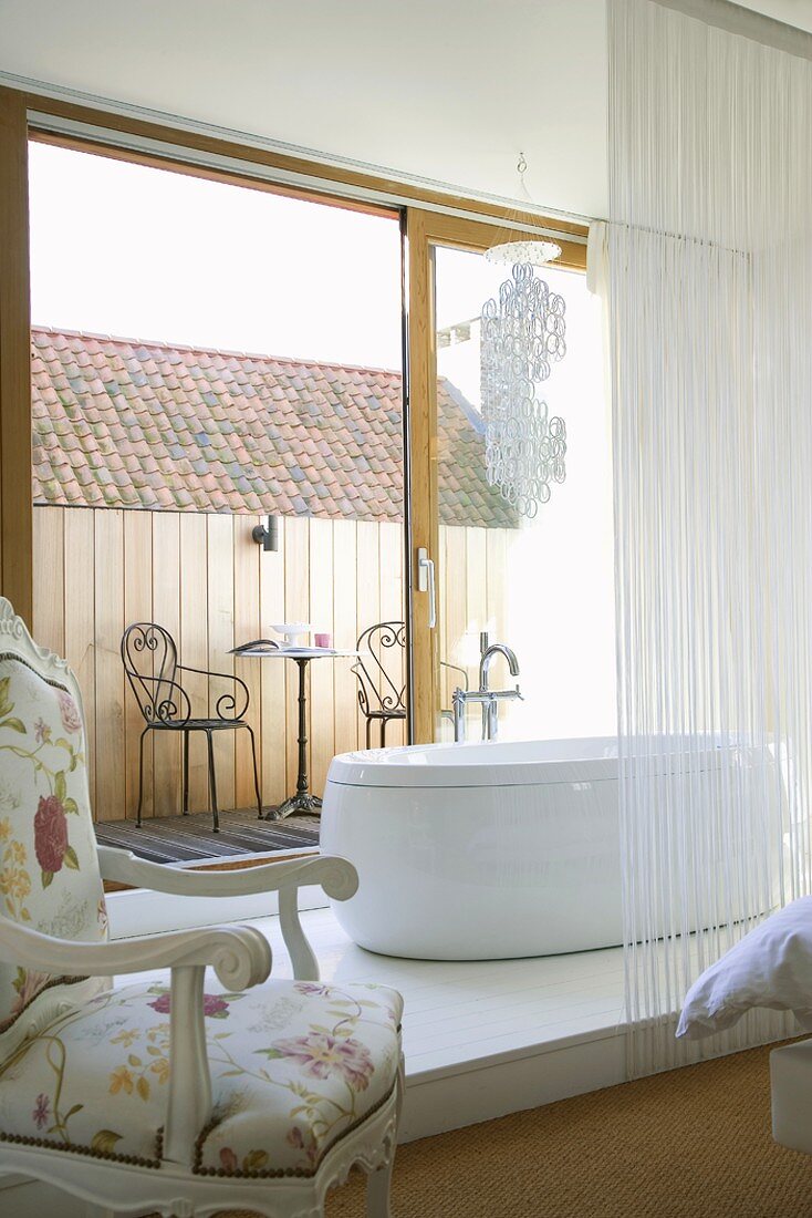 Moderne, freistehende Badewanne auf einem Holzpodest vor der geöffneten Balkontür im Schlafzimmer