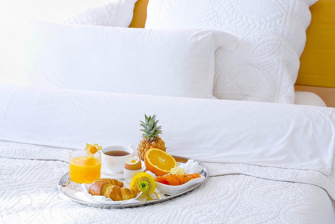 Frühstückstablett mit frischen Früchten, Croissant und Tasse Tee auf weißbezogenes Bett im Hotelzimmer