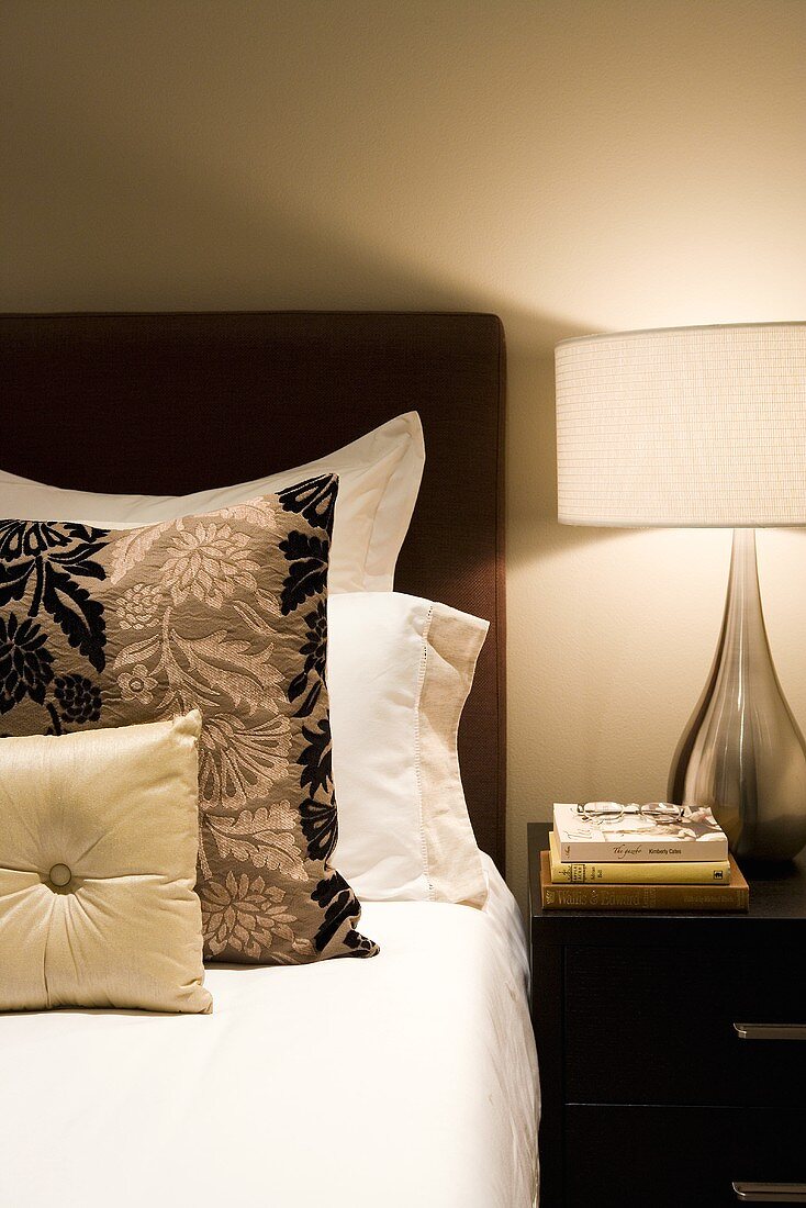 Bett mit verschiedenen Zierkissen und Nachttisch mit Lampe & Büchern im Schlafzimmer (Ausschnitt)