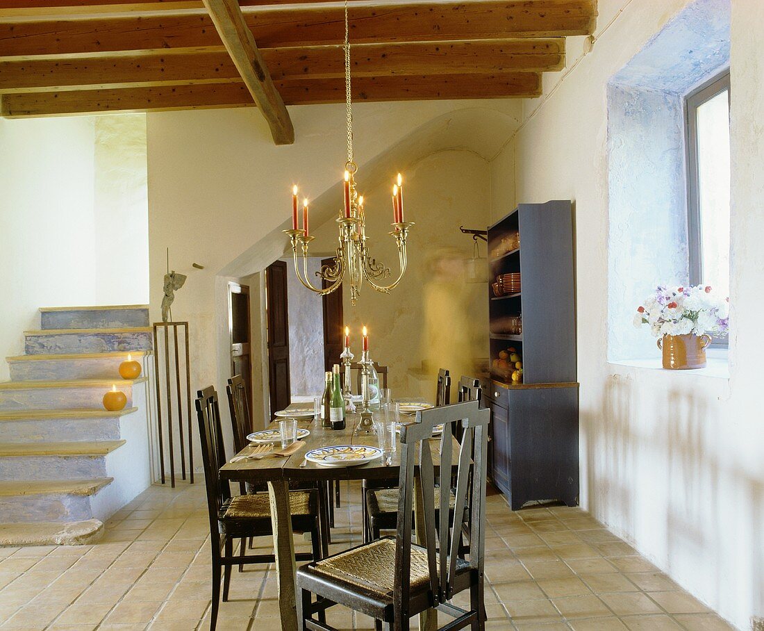Ländlicher Wohnraum mit Balkendecke, gedecktem Tisch, hängendem Kerzenleuchter & Treppenaufgang aus Stein