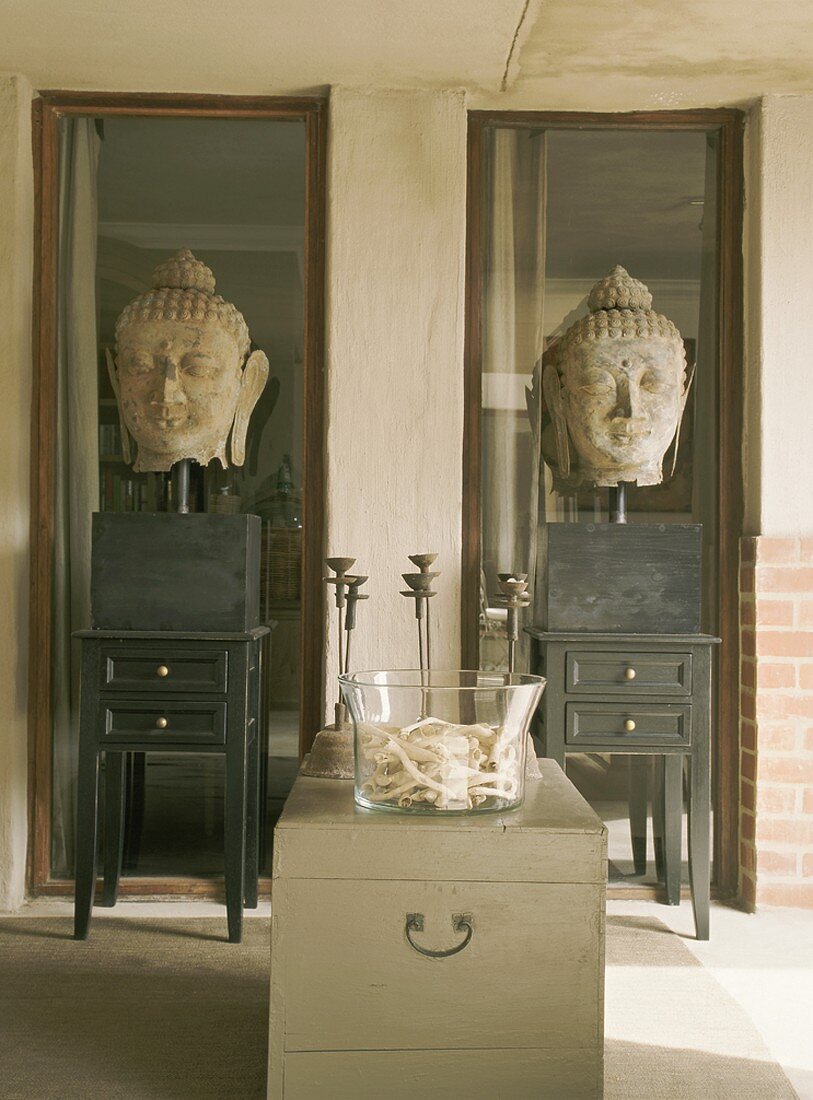 Zwei steinerne Buddha-Köpfe auf Tischchen vor deckenhohen, schmalen Fenstern