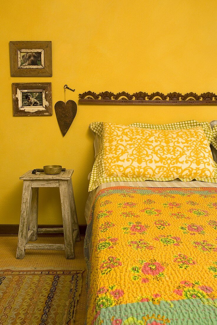 Bed with a printed bedspread and rustic stool in front of a yellow wall
