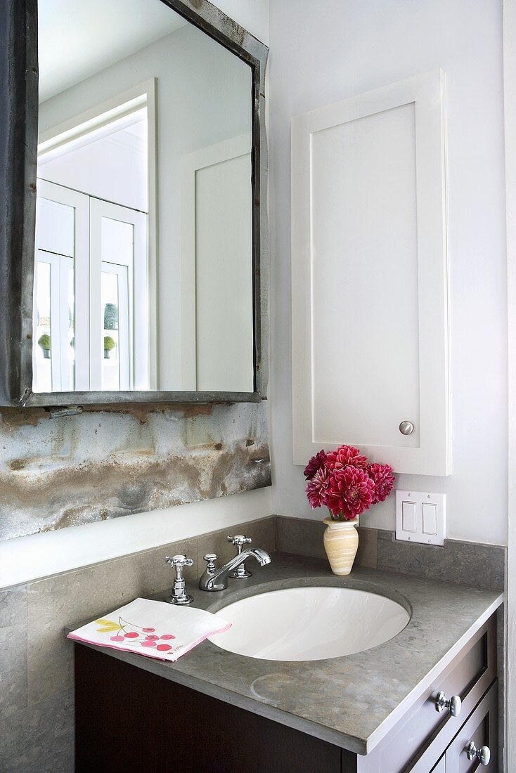 Eleganter Waschtisch mit rotem Blumensträusschen unter derbem Badezimmerspiegel mit geschweisstem Metallrahmen