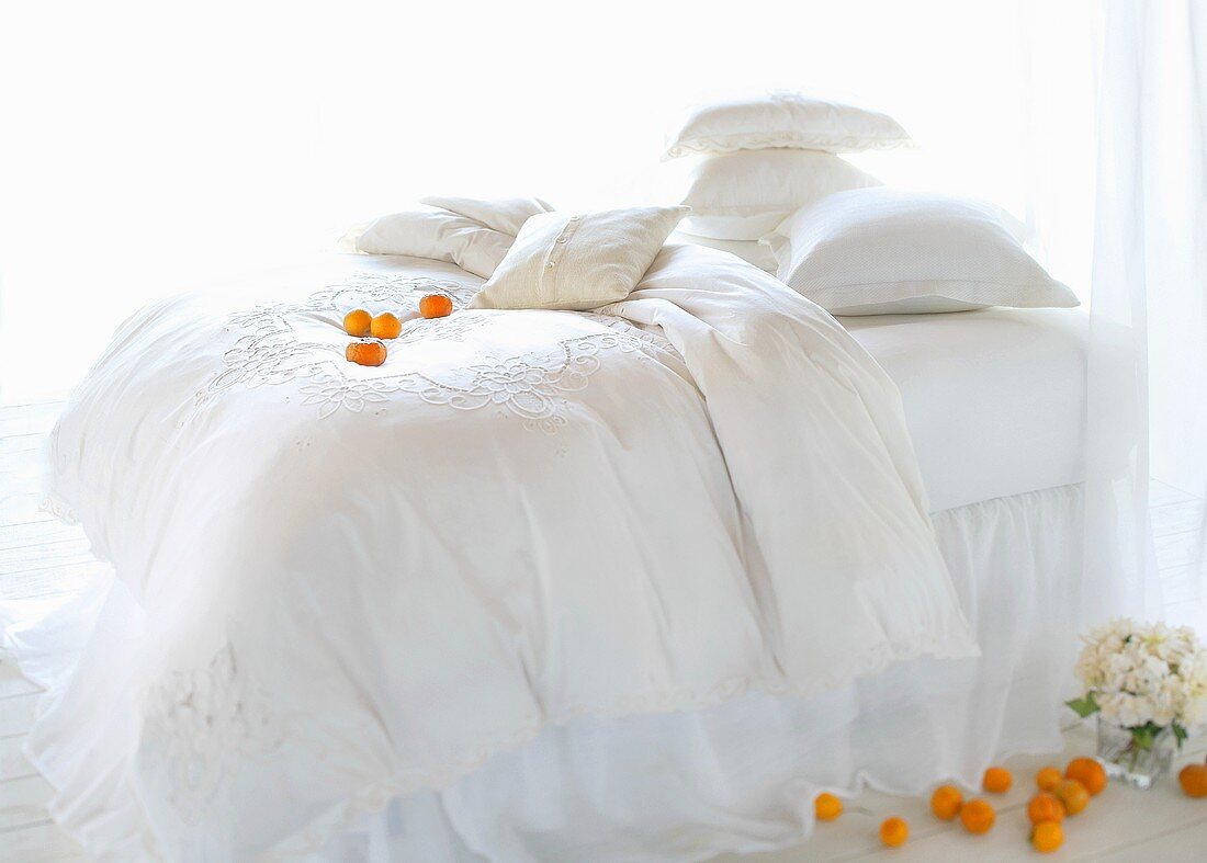 Himmlisch weiches Bett in Weiß mit vielen Kissen und bestickter Bettdecke, daneben ein Blumenstrauss und Mandarinen