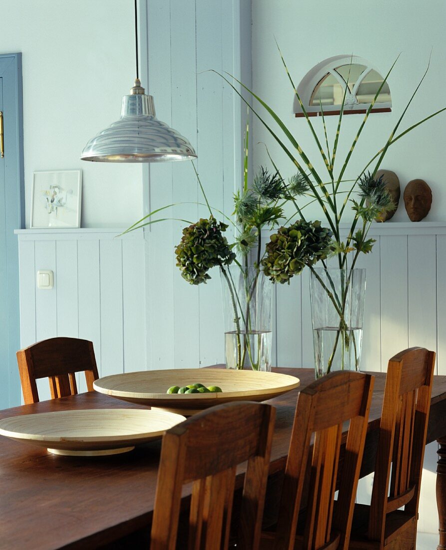 Brauner Esstisch aus Massivholz mit passenden Stühlen; darauf große Holzschalen und zwei Vasen mit getrockneten Hortensienblüten