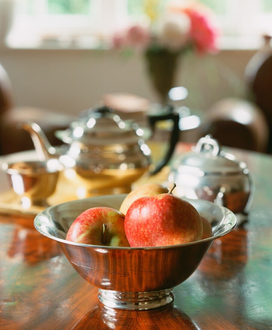 Äpfel in einer Silberschale, im Hintergrund Teekanne