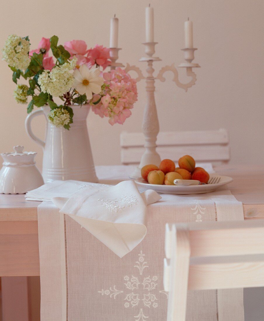 A bunch of flowers in a jug, a candle holder and a plate of fruit