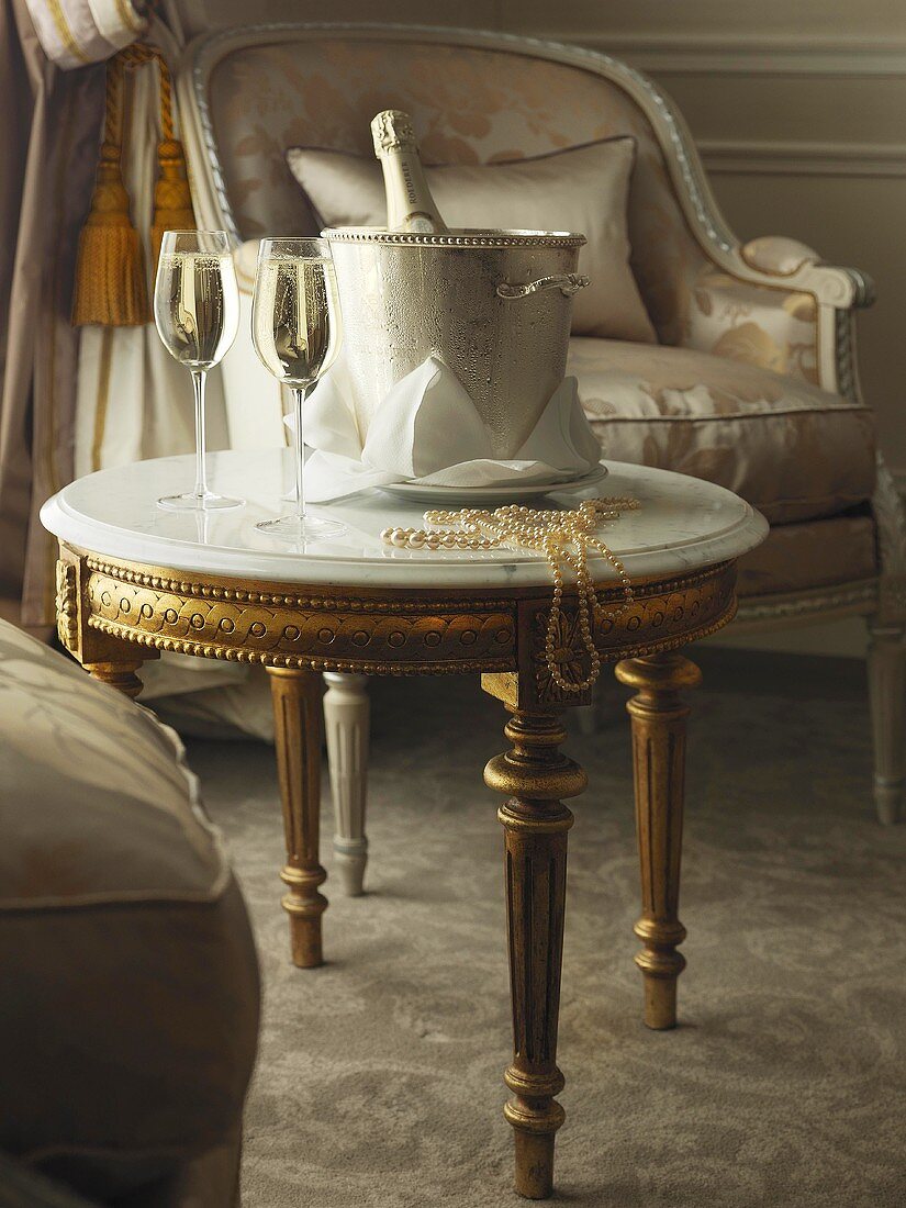 Vergoldeter, runder Beistelltisch mit Marmorplatte in vornehmem Ambiente; darauf ein silberner Sektkühler und perlender Sekt in Gläsern
