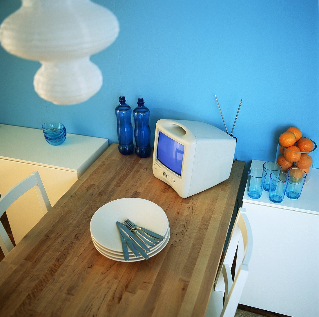 Esstisch aus Holz vor einer blauen Wand mit Tellerstapel, Besteck und einem Minifernseher