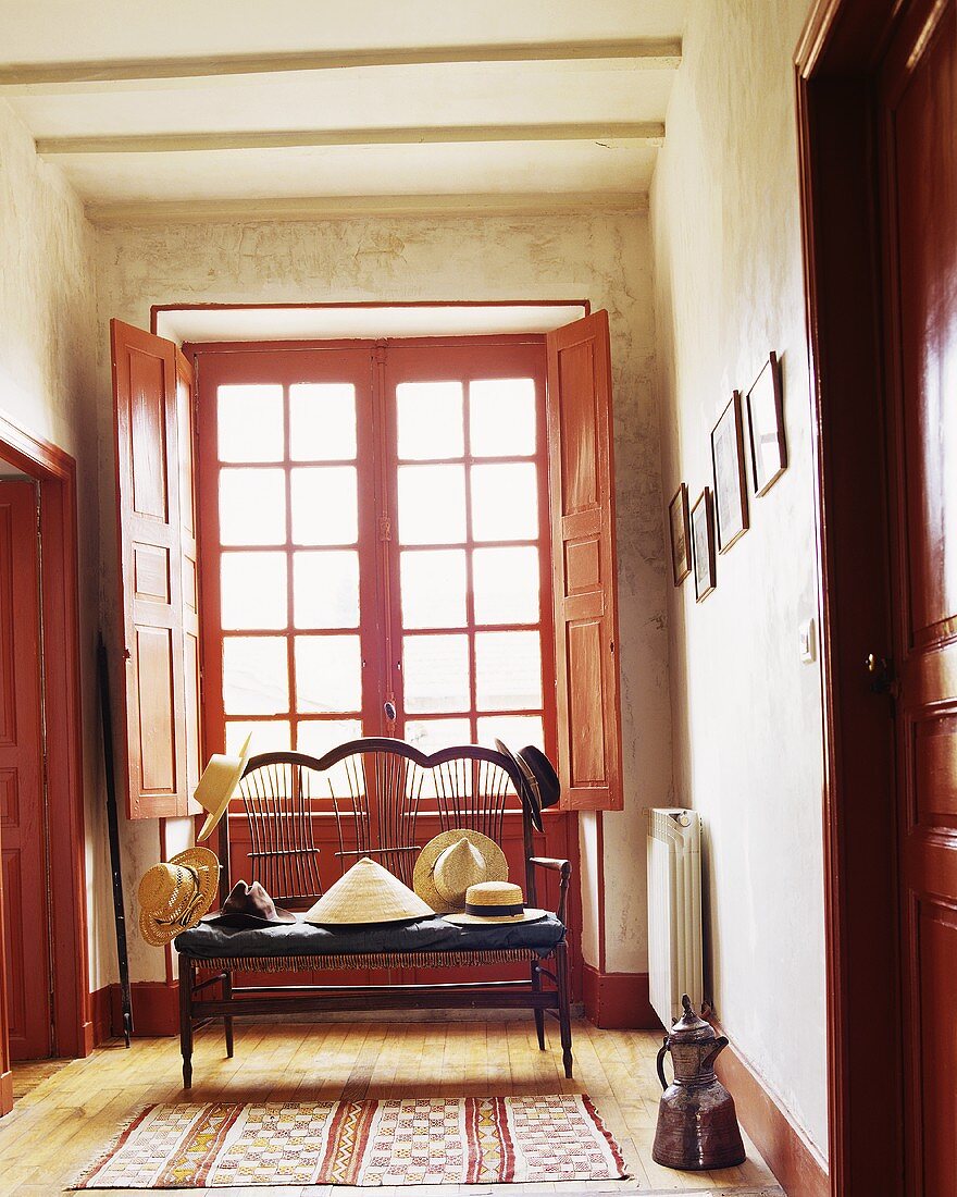 A window with a red frame in a corridor with various hats on a seat in front of it