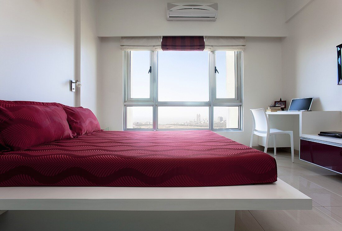 A bed with red bed clothes in a white room with a large window