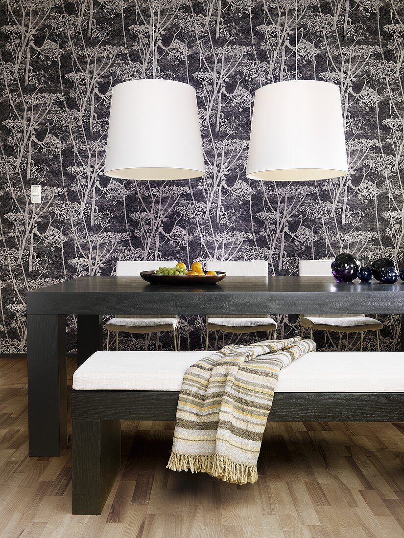 White lampshades hanging above a table and an upholstered bench against a black and white papered wall