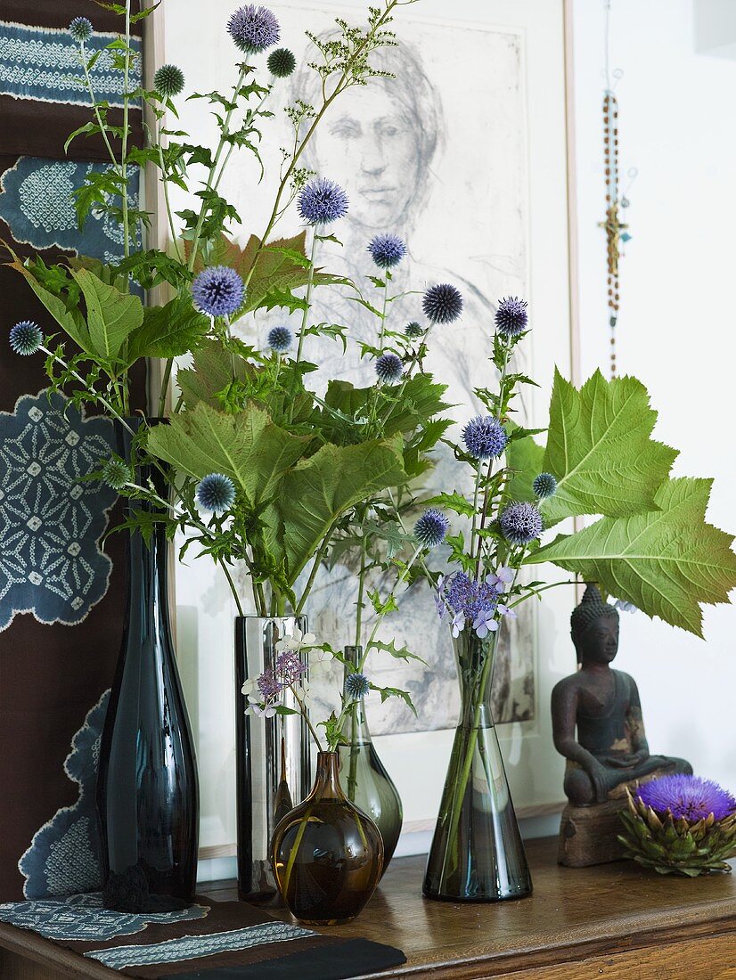 Purple flowers in assorted vases next to a statue of the Buddha on a wooden shelf