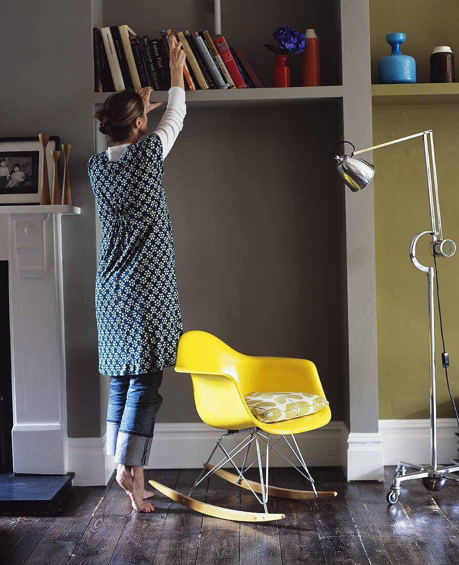 Wohnraum mit gelbem Schaukelstuhl aus Plastik, Stehlampe auf Rollen und einer Frau vor Bücherregal