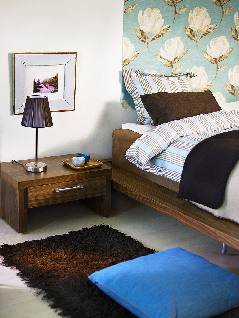 Schlafraumecke mit Nachttisch aus dunklem Holz und Wandbehang mit Blumenmuster vor Bett