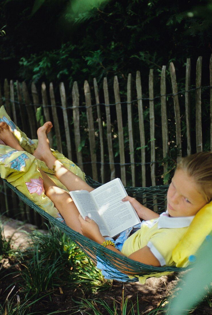 A girl lying in a hammock reading a book in a garden