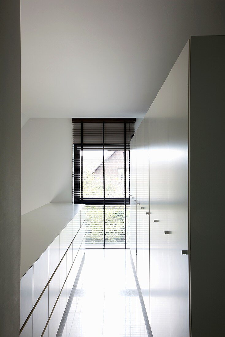 A narrow room with a white sideboard and a tall white cupboard on the long side with a floor-length window at the end