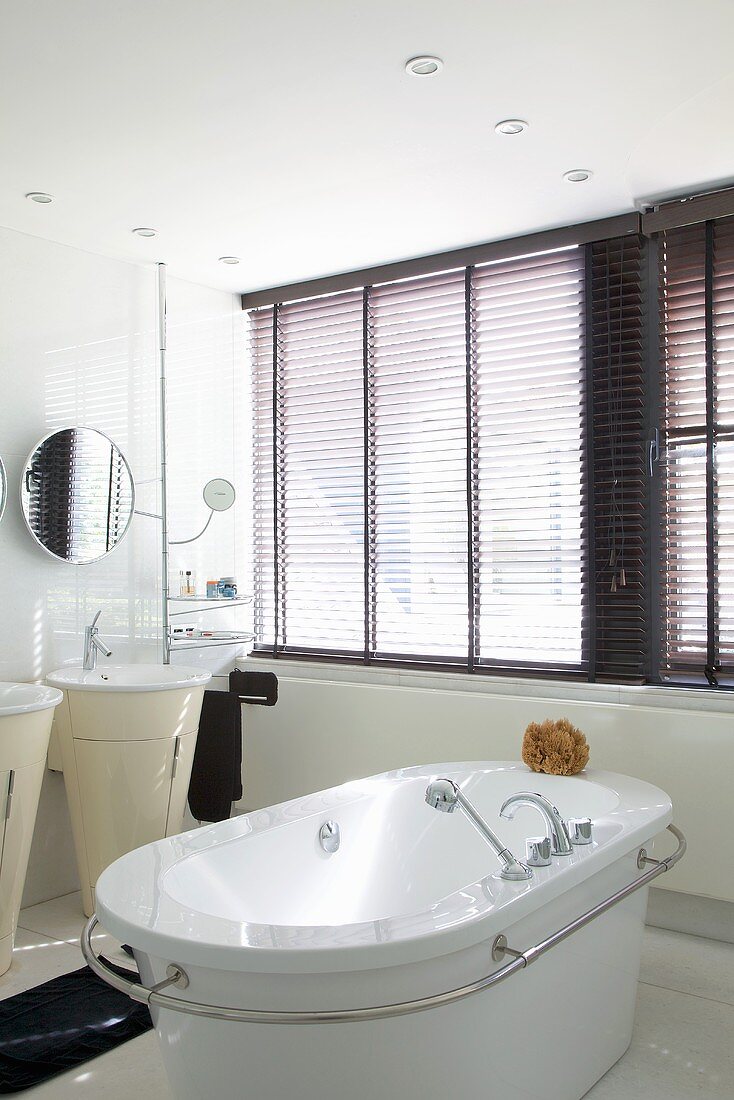 A free-standing bathtub with a circumferential stainless steel bar in a white bathroom with a large window bank