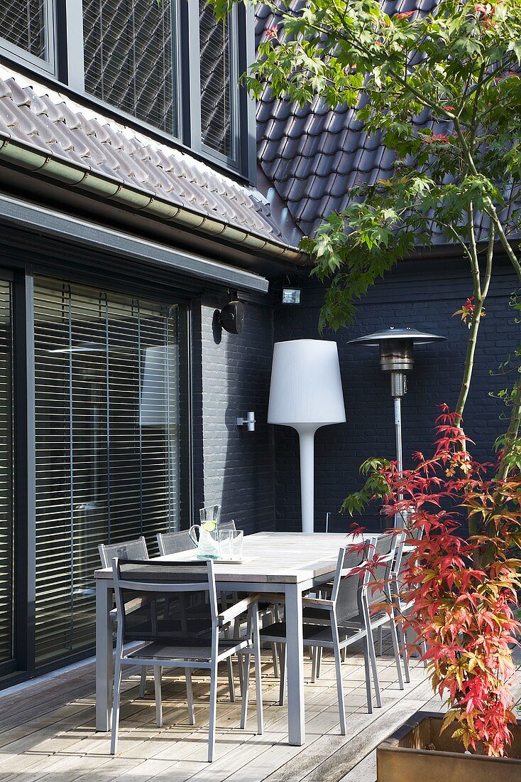 A terrace in the corner of a house furnished with a table, chairs and a floor lamp