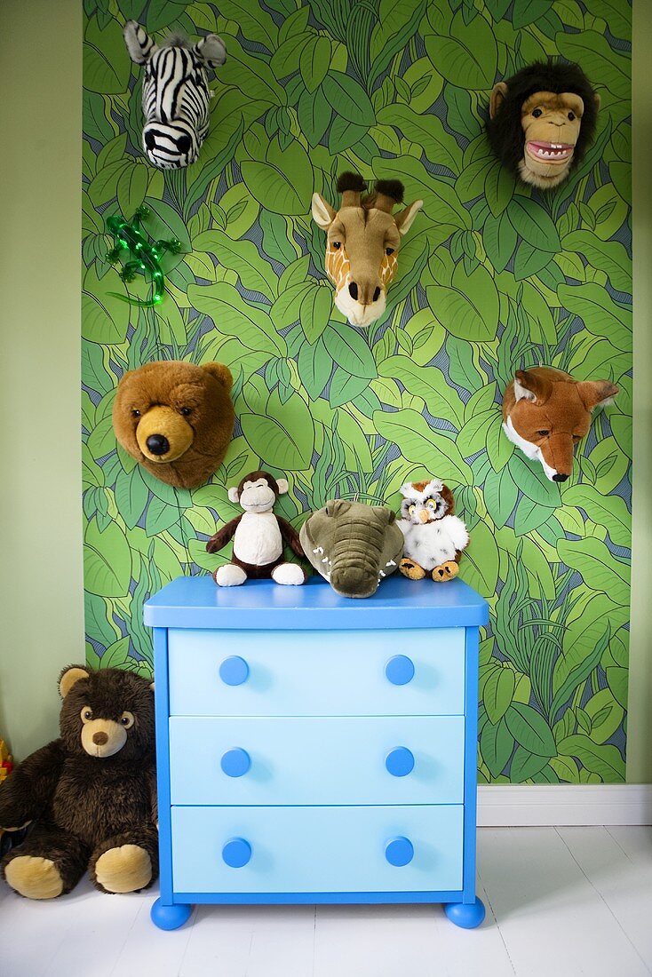 A child's room - a blue chest of drawers in front of a wall with painted jungle design and hung with plush animal heads
