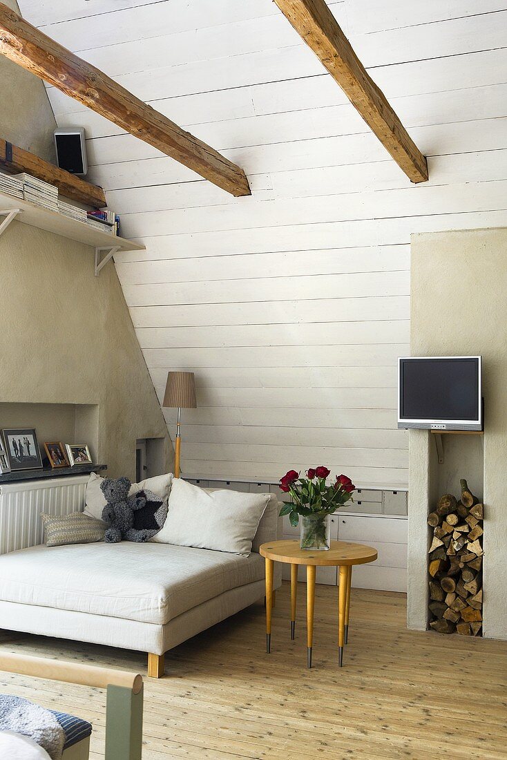 Bett und Beistelltisch unter Dachschräge mit weisser Holzverkleidung und TV über Holzstapel an Betonwand