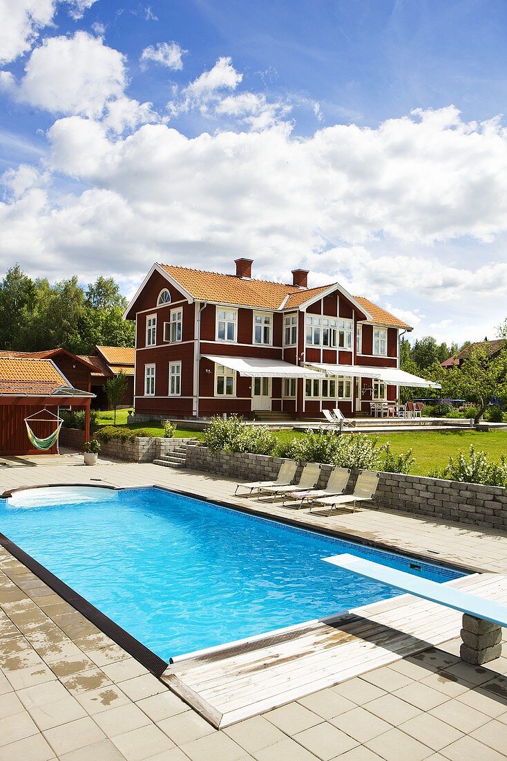 Pool mit Sprungbrett und rotbraunes Holzhaushaus mit weissen Fenstern